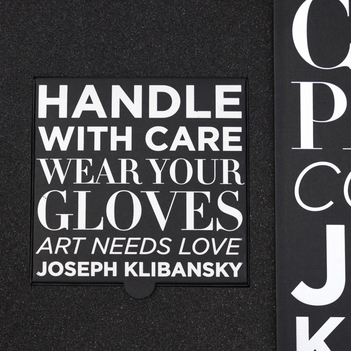 Black box with white text 'HANDLE WITH CARE WEAR YOUR GLOVES ART NEEDS LOVE JOSEPH KLIBANSKY' on a black background.