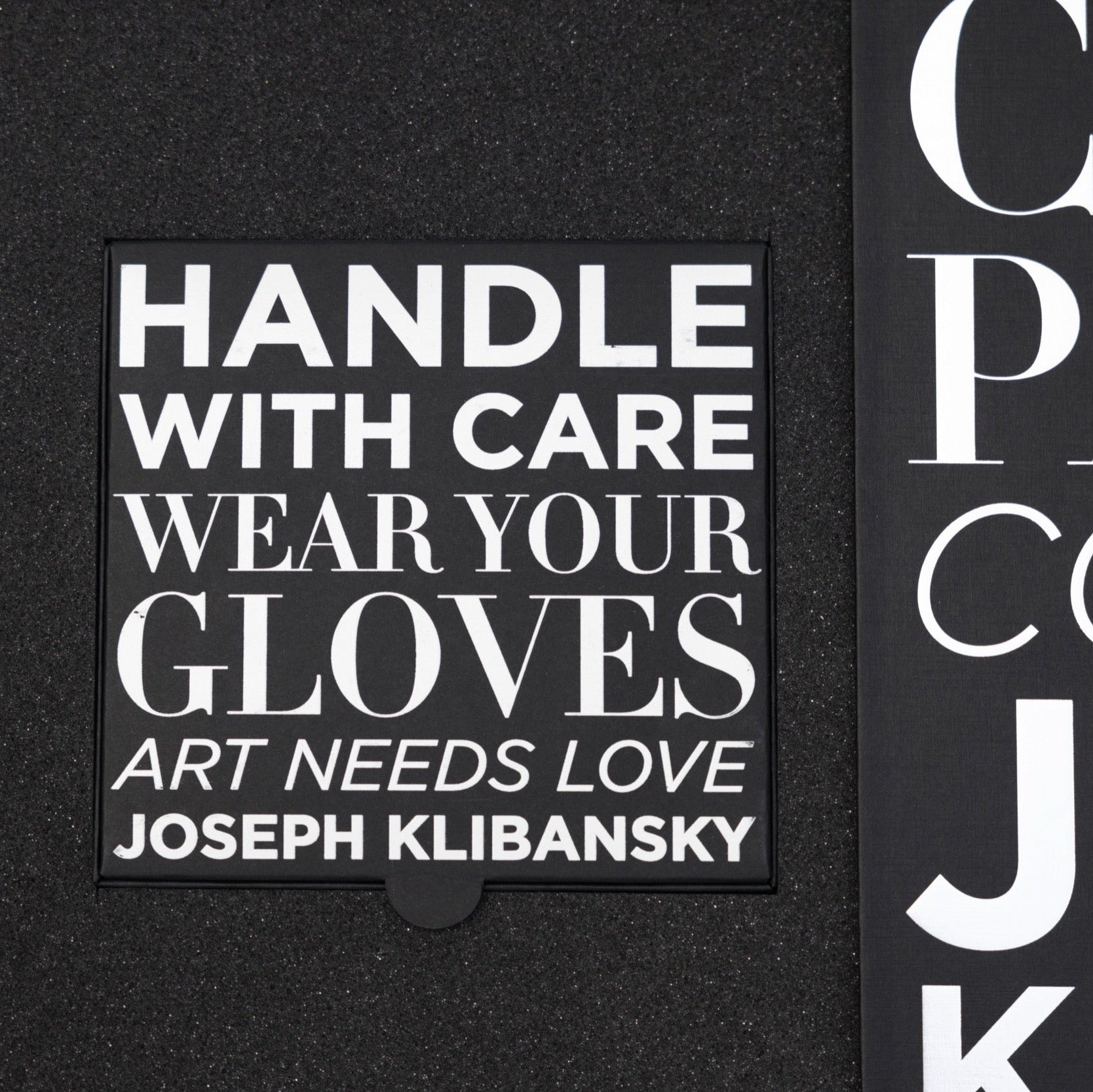 Black box with white text 'HANDLE WITH CARE WEAR YOUR GLOVES ART NEEDS LOVE JOSEPH KLIBANSKY' on a black background.