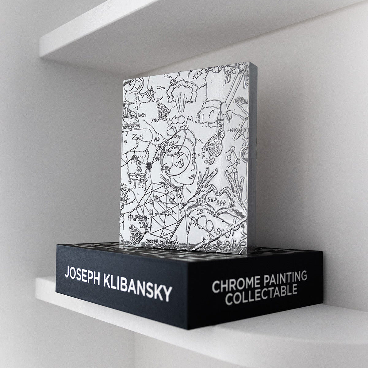 A chrome painting collectable art collectable by artist Joseph Klibansky standing on a black box on a white shelf from a slight angle