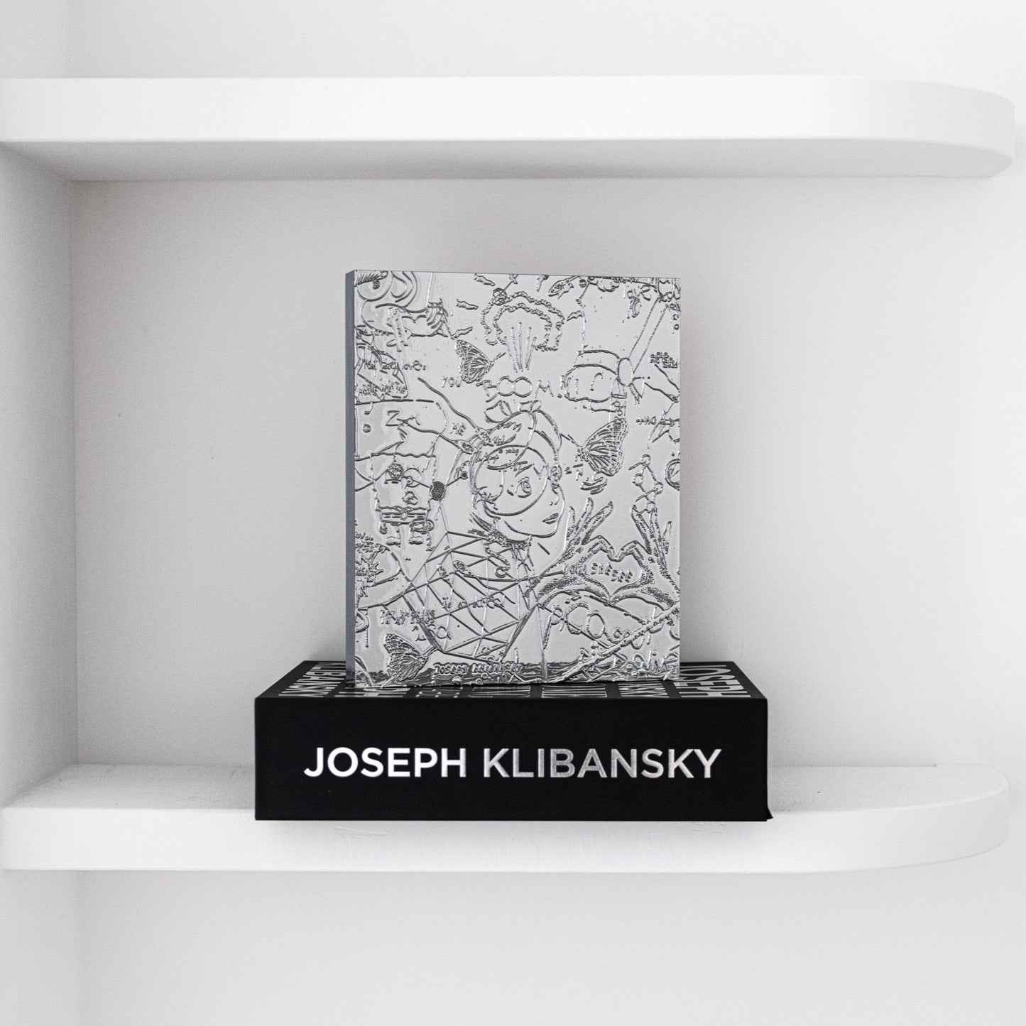 A chrome painting collectable art collectable by artist Joseph Klibansky standing on a black box on a white shelf