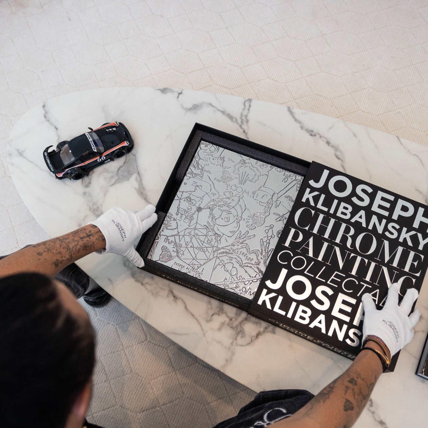 Person holding a chrome painting collectable art collectable by Joseph Klibansky in a black box on a marble coffee table with a scale model of a Porsche car next to it
