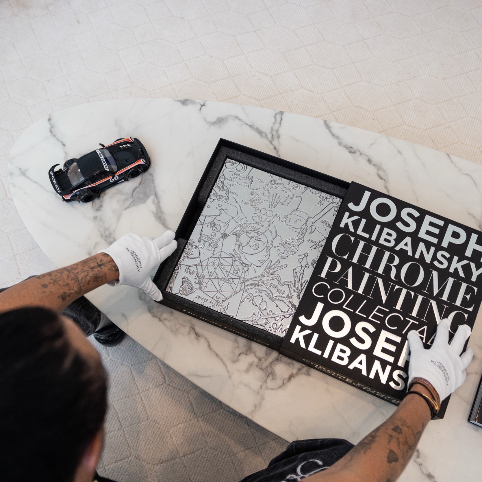 Person holding a chrome painting collectable art collectable by Joseph Klibansky in a black box on a marble coffee table with a scale model of a Porsche car next to it
