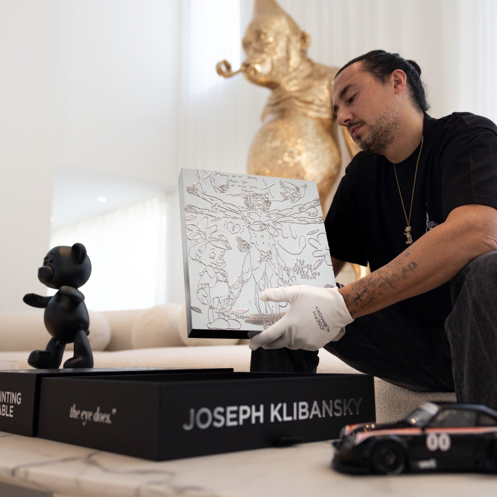 Man in a black shirt and white gloves holding a framed artwork next to a black bear figurine and a black box with 'JOSEPH KLIBANSKY' on it.