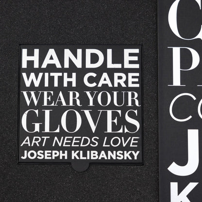 Black box with white text 'HANDLE WITH CARE WEAR YOUR GLOVES ART NEEDS LOVE JOSEPH KLIBANSKY' on a black background.